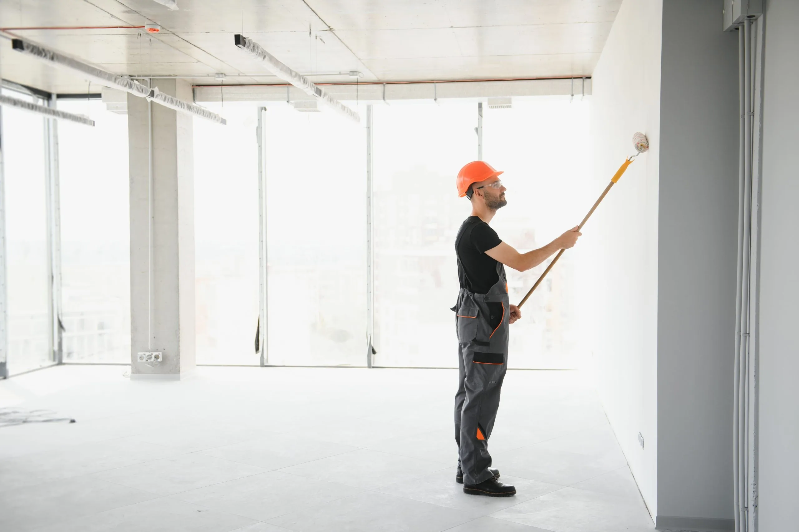 Professional commercial painter in orange hardhat rolling white wall in large office space by Bos-Flo Coatings