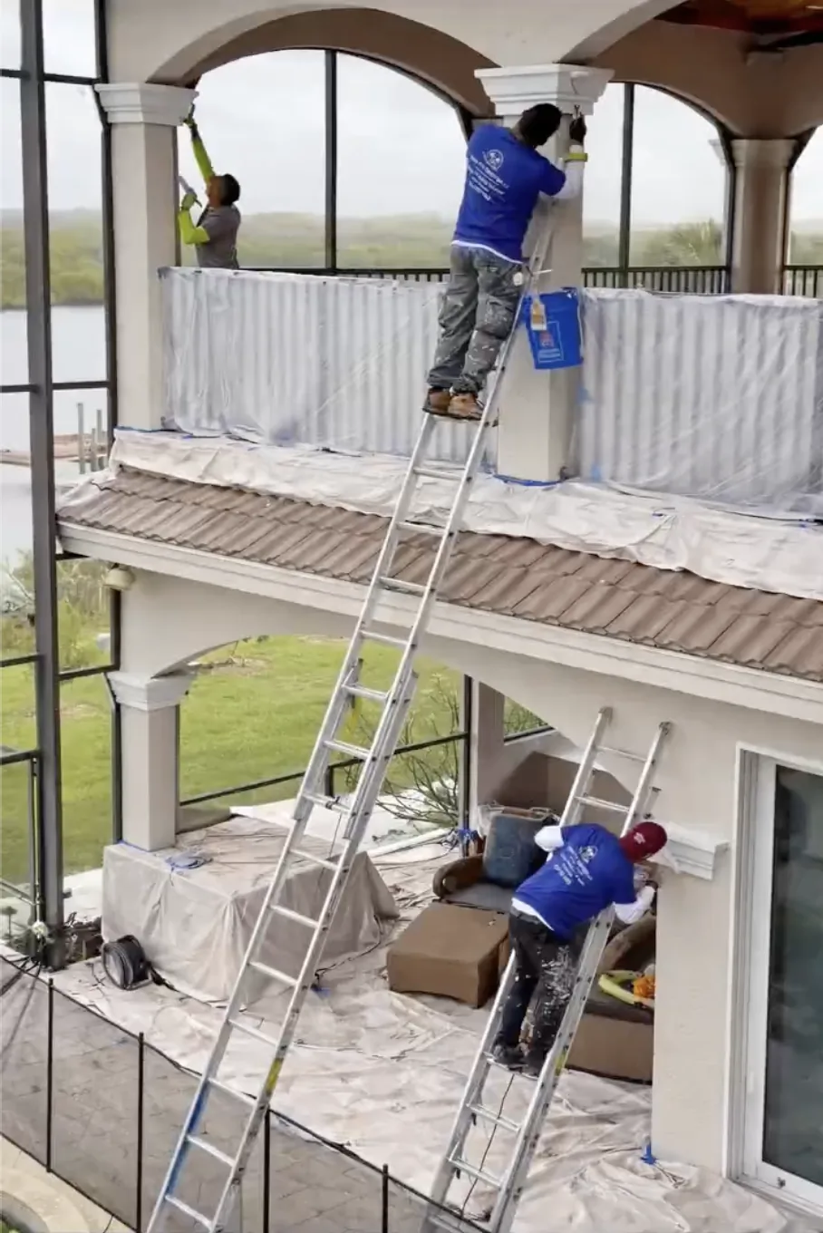 Painters on ladders applying exterior paint to pillars and trim of a two-story home with terrace and outdoor seating area